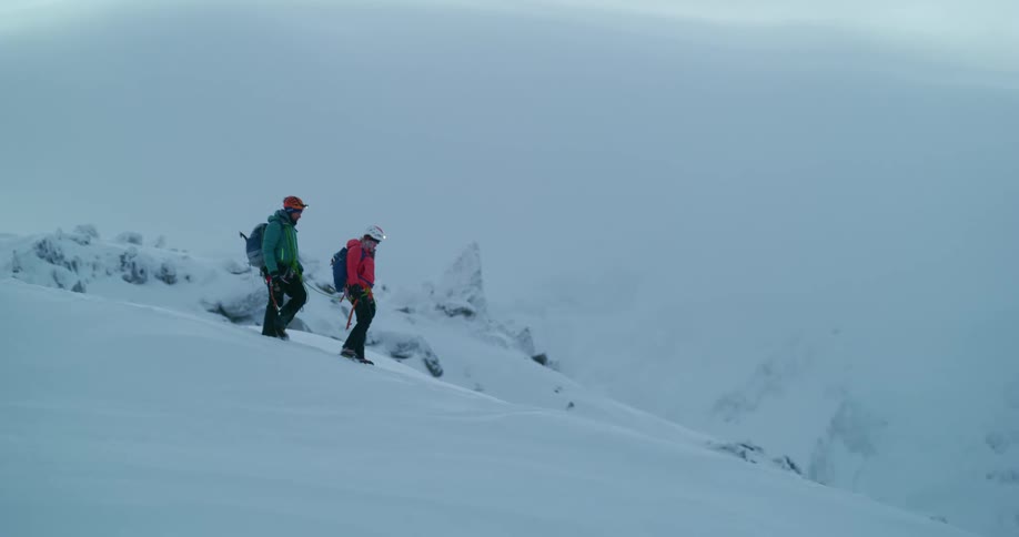 Two people tied together with safety rope hike down shallow slope in snow covered mountains during winter