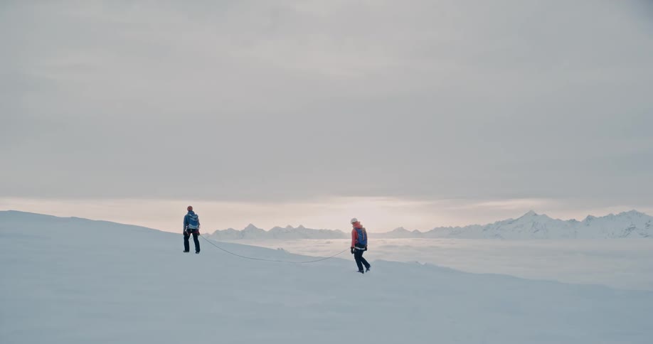 Two hikers walk across a snow covered mountain terrain as the sun is setting beyond the horizon