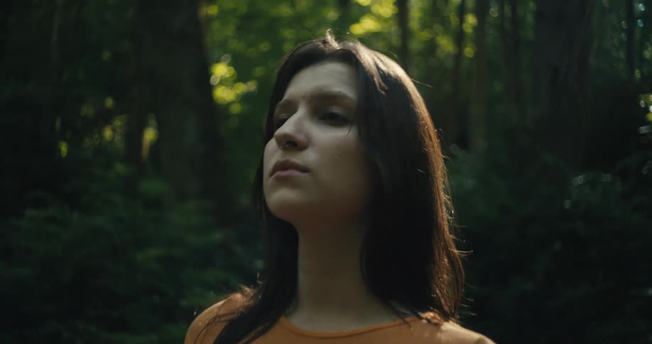 A woman with a nose piercing wearing an orange tshirt stands in the forest closing her eyes and gazing upwards in quiet contemplation