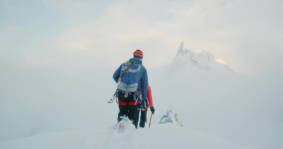 Hikers in snow gear track across a snowy mountain terrain against a white snow covered background peak and sky