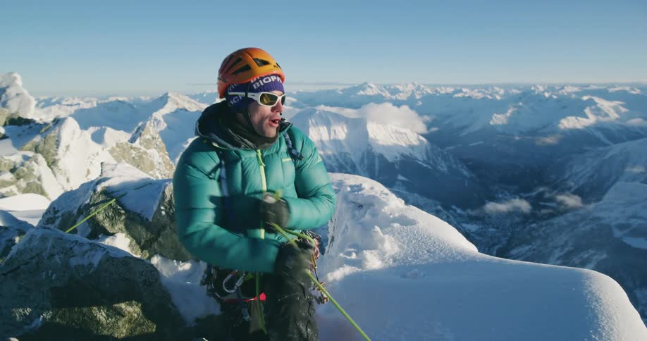 A mountain climber on the peak of a snow covered mountain pulls his rope as he climbs overlooking mountains in the distance