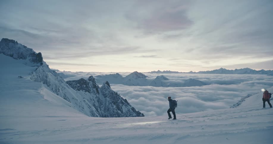 Two people wearing headlamps walk across a snow covered mountain high in the clouds with mountains in the distance