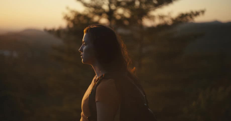 Female hiker stands overlooking forested valley under a red tinted sky on a windy day