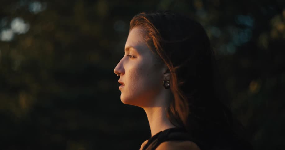 The sun shines on womans face with a nose piercing and earrings near the trees waving in the wind