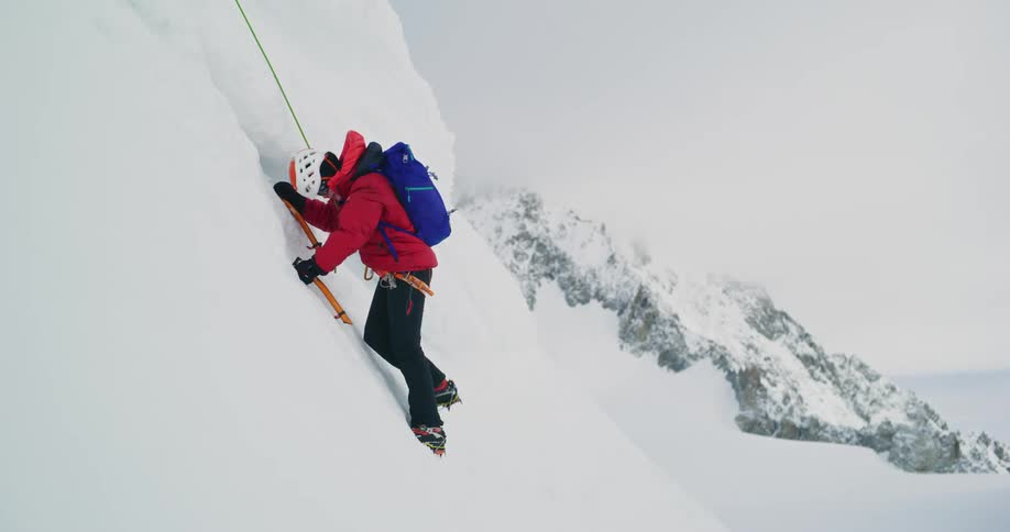 A man wearing a backpack holds his trekking poles as he climbs a steep snow covered mountain slope