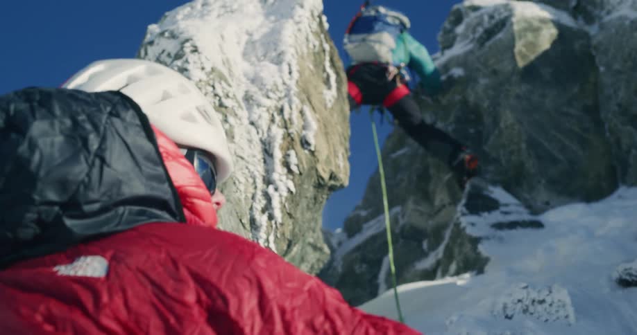 A mountain climber stands on a rock while another climber waits below on a winter day