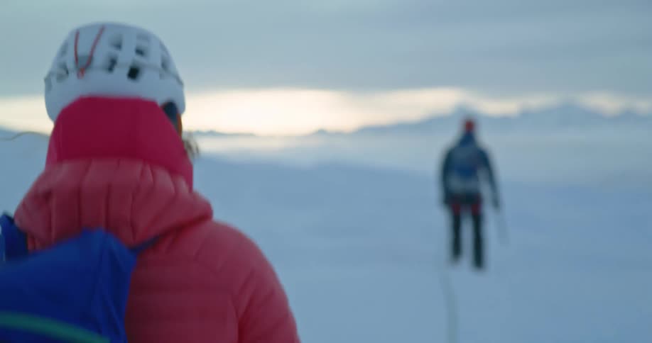 Female hiker stops behind partner and looks around a snowy terrain as the sun is setting
