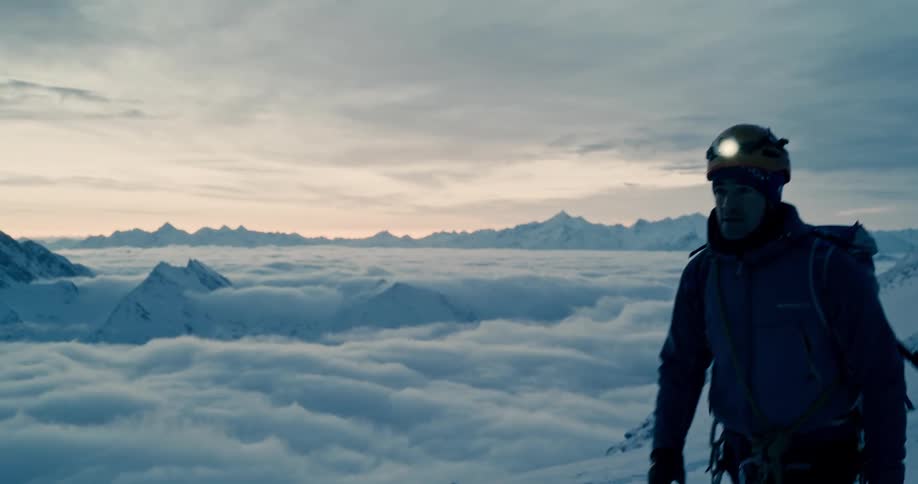 A man wearing a headlamp walks across a snow covered mountain ledge high in the clouds with mountains in the distance