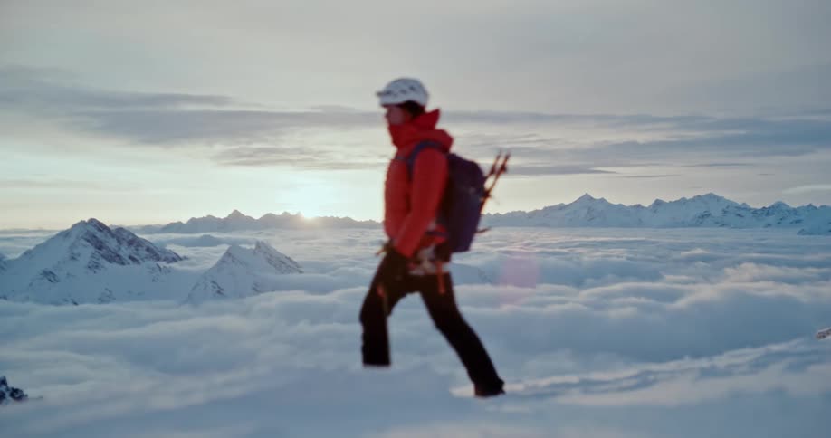 Female mountaineer walks through snow wearing extreme weather winter gear with white peaks in background at sunrise