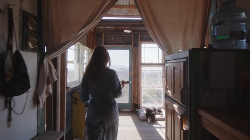 Woman carries a plate of food through a small home in the desert towards the front door