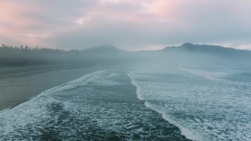 Waves roll into shore and over the beach with mountains in the distance under a cloudy sky