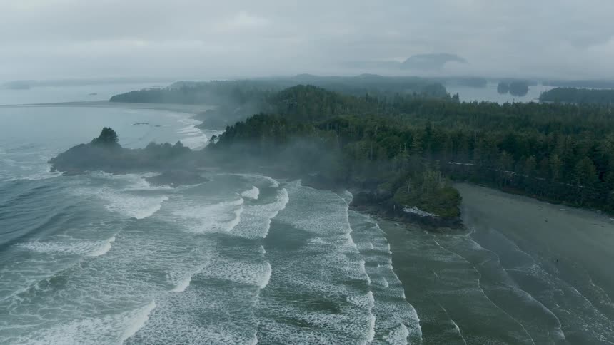 An aerial view capturing ocean waves breaking against a sandy beach and rocky shore