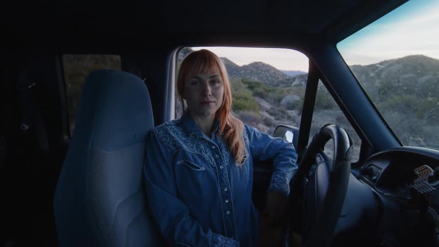 A woman is sitting in the drivers seat of a parked truck looking ahead then turning to look out of the side window