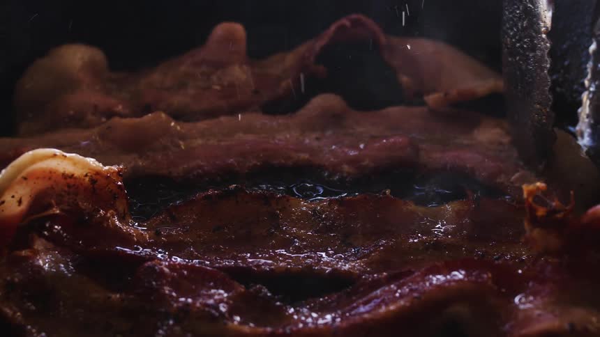 Tongs are moving and flipping strips of bacon frying in a pan