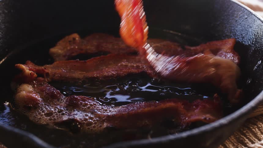 Tongs flip bacon frying in a pan