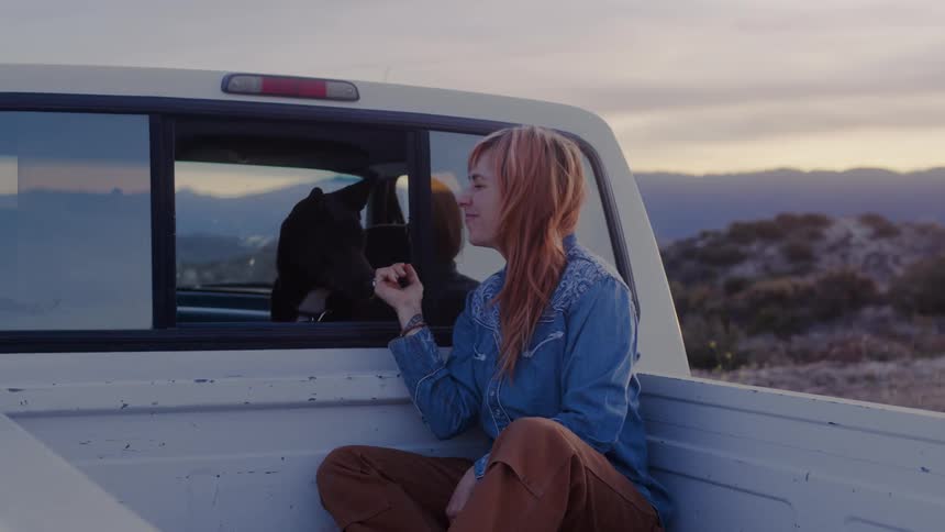 Teen seated at the back of a pickup truck pet a dog at sunset