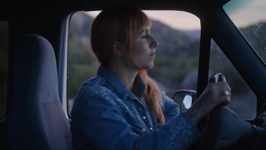 A woman is sitting in the drivers seat of a truck holding the steering wheel looking out of the side window and then turning her head looking in front