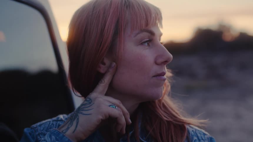 A woman with red hair a blue tshirt and a scorpion tattoo on her hand leaning against a pickup truck