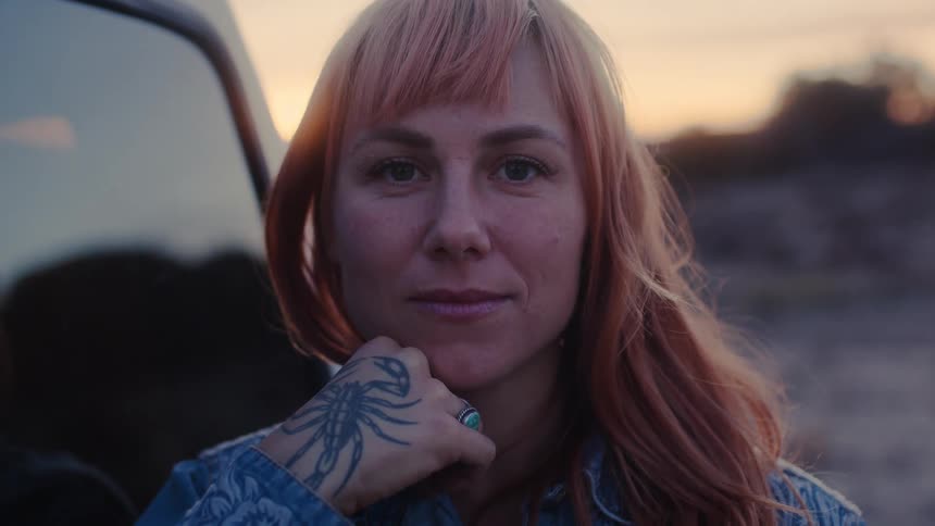 Portrait of a woman with a jacket and hand tattoo in an outdoor setting with a reddish sky