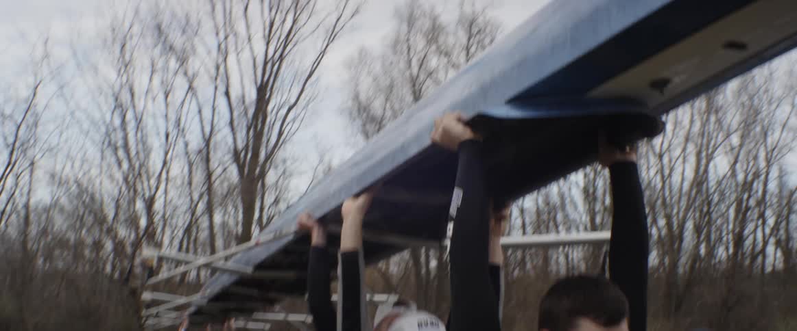 A male row team holds a boat above their head and carries it towards the water