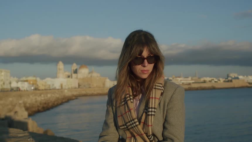 A woman in cold clothes is wearing sunglasses and behind her is a city facing the sea