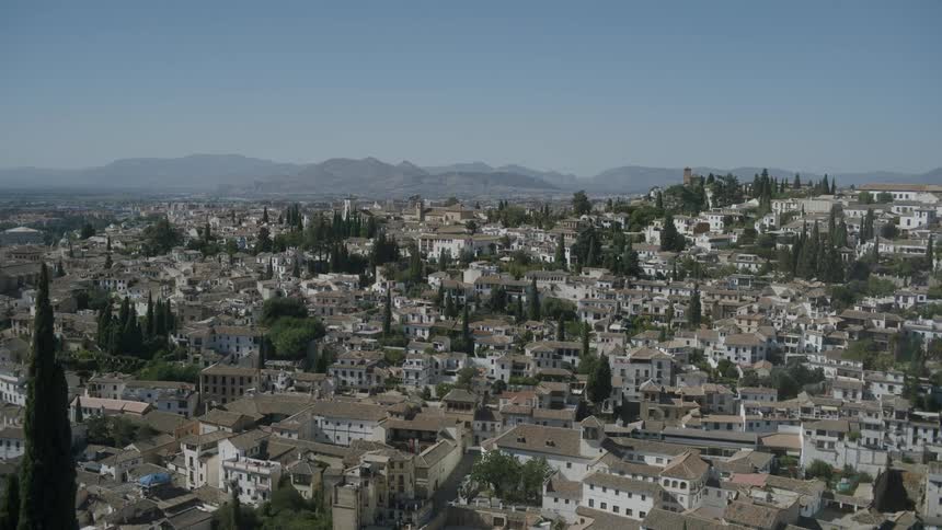 Tower cam view of white residential houses with gray roofs dotted with tall trees on a spanish hillside during the day