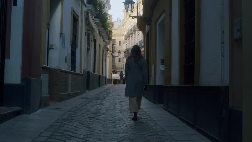 Back profile of a female with brown hair wearing a long blue coat and trousers walking on a shadowed narrow cobblestone road through apartment buildings with spanish style facade on a sunny day