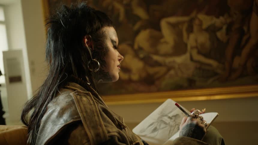A woman with tattoos and earrings is drawing something with paper and a pencil and is looking straight ahead