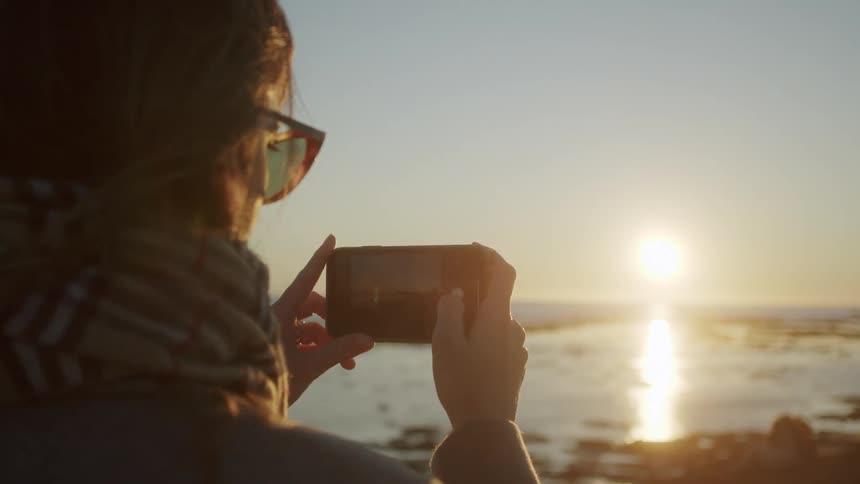 Persona wearing sunglasses and scarf takes smartphone photo of sun over sea