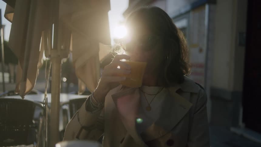 A woman wearing sunglasses is sitting and having a drink while the sun