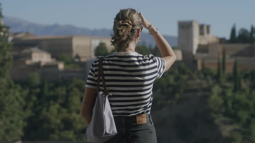 A woman with her back turned facing a view of monasteries amidst mountains and vegetation in the daylight of a cloudless blue sky