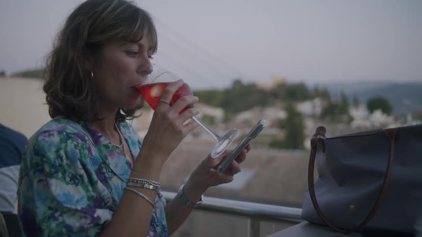 Woman sitting outdoors in scenic town drinks wine while scrolling on her smartphone