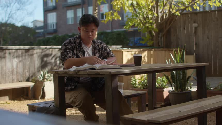 Young man sitting at an outdoor table writing and reading a book