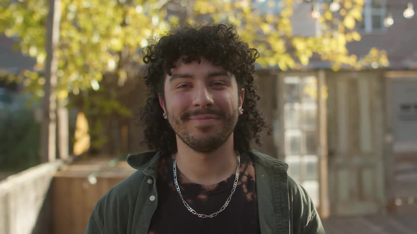 Man with curly hair a beard a necklace and earrings smiles at camera in the yard