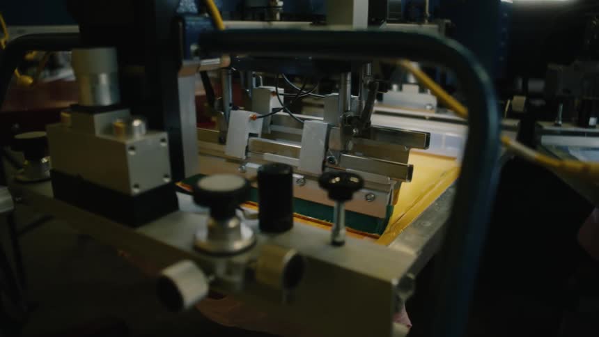 Scrapper pulls yellow ink across the tray with a tee shirt underneath of an automated screen printing machine in a screen print factory