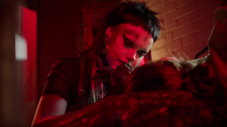 Heavily tattooed tattoo artist works on a client who is lying down in a dimly lit room