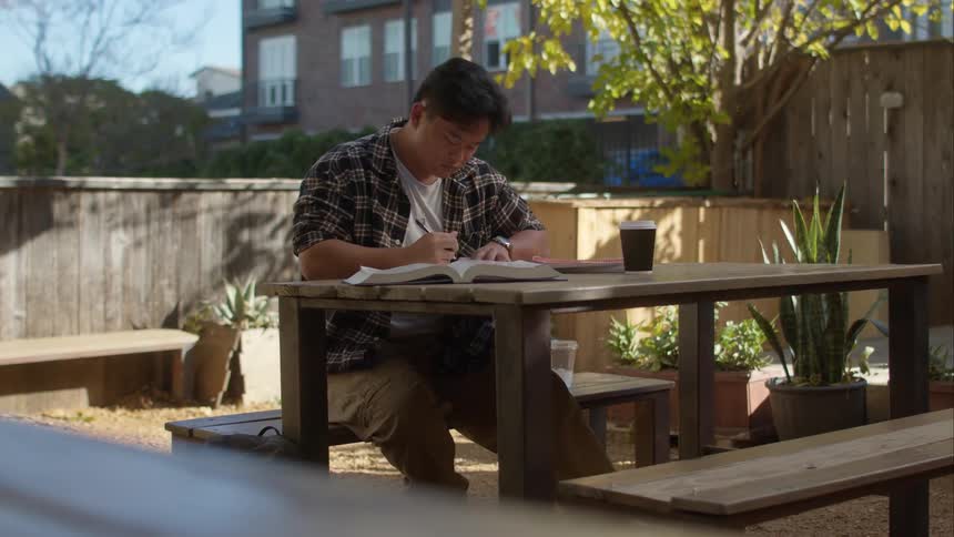 A teenage boy is sitting outside at a table writing and doing his homework