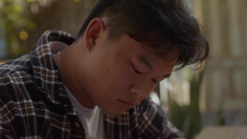 A teenage boy is sitting at a table looking at a book and writing in a notebook