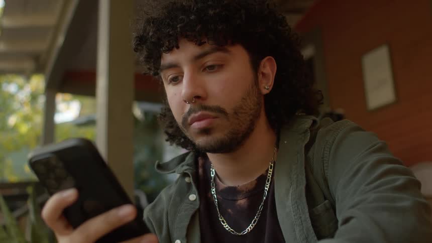 A man with a nose piercing beard earrings and a necklace using a smartphone while sitting on the porch