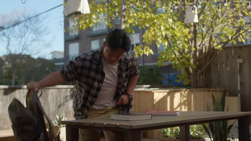 A young man approaches a table and takes books from his backpack and sits down to study them