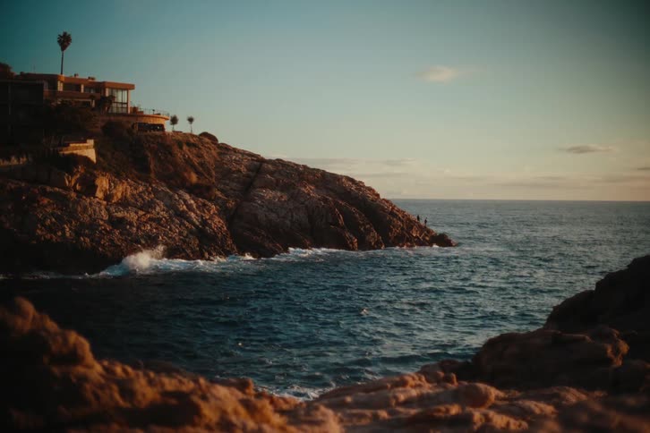 Waves crash into the rocks of a cove below a california beach house with rocks reflecting golden light at sunset with a clear sky and ocean in the background