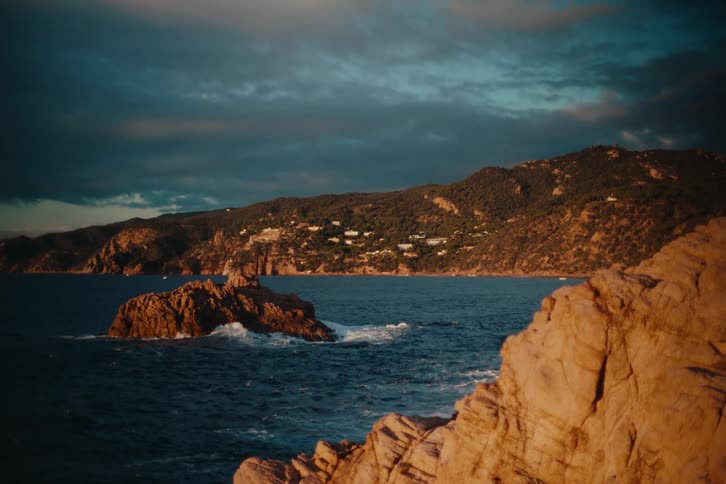 Waves splash against rocks along the coast with buildings on the mountainside overlooking the water