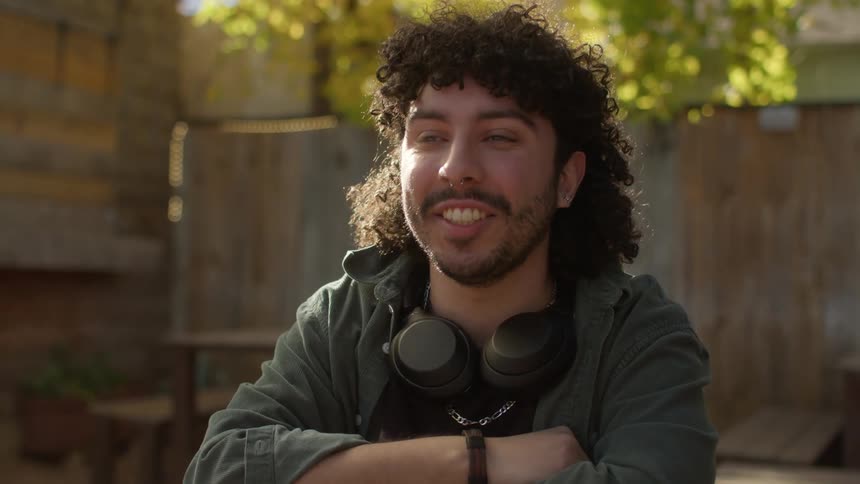 A man with headphones around his neck is sitting outside smiling and nodding his head in agreement