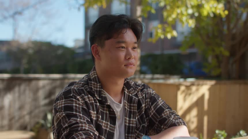 A teenager in a plaid shirt sits at a table sipping a beverage from a plastic cup in a cafe yard