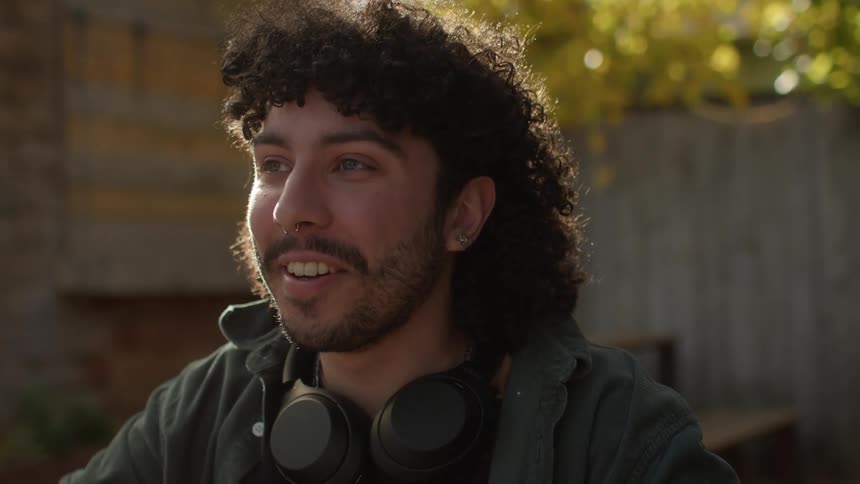 A man with curly hair earrings and headphones sitting at a table smiling in the yard