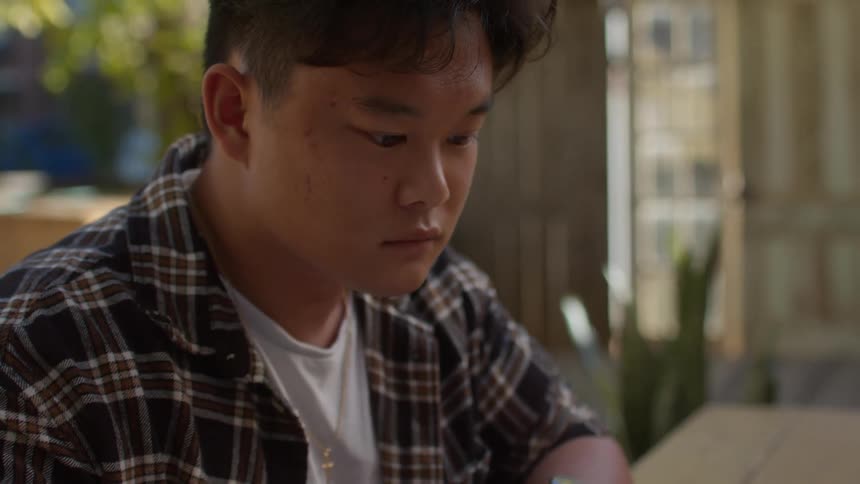 A teenage boy is sitting at a table looking at a book and writing in a notebook