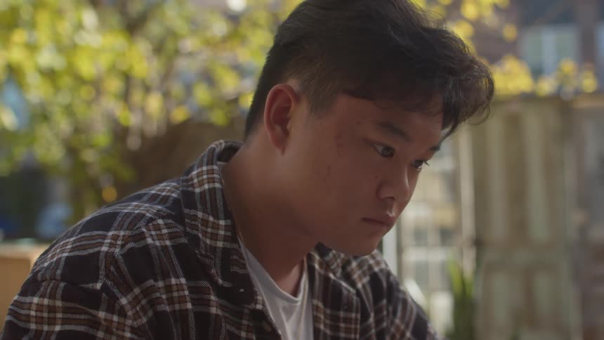 A teenager in a plaid shirt and white tshirt sits at a table writing in a notebook in the yard