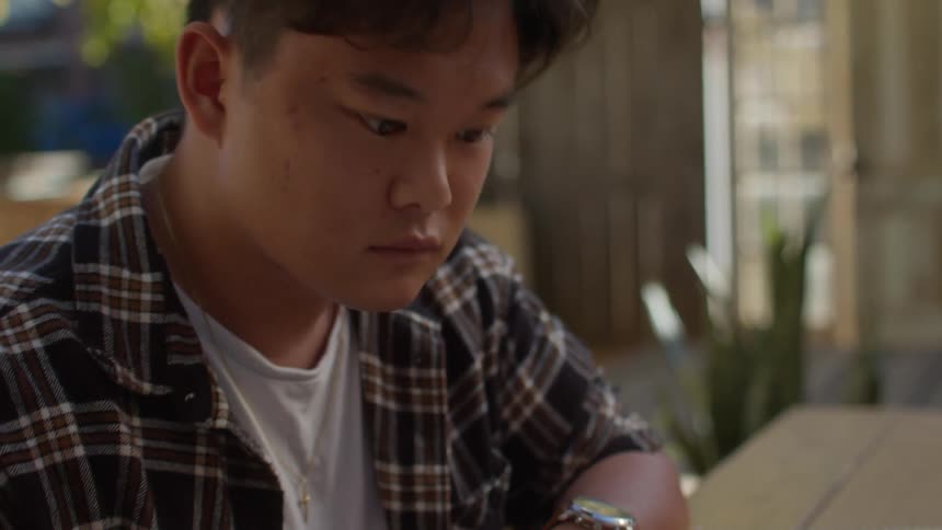 A teenage boy is sitting at a table looking at a book and writing in a notebook