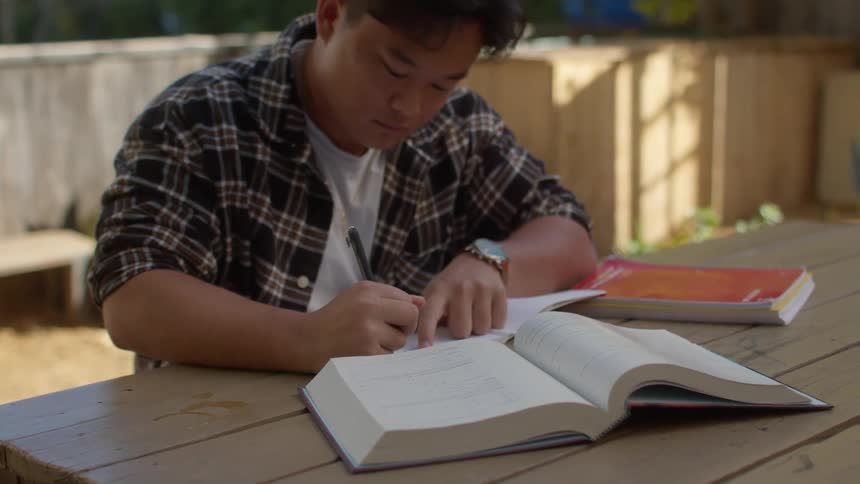 Young man sitting at a wooden table in the backyard writing while reading a book