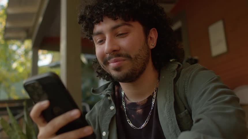 A man is sitting on a porch holding his cell phone and smiles as he nods his head